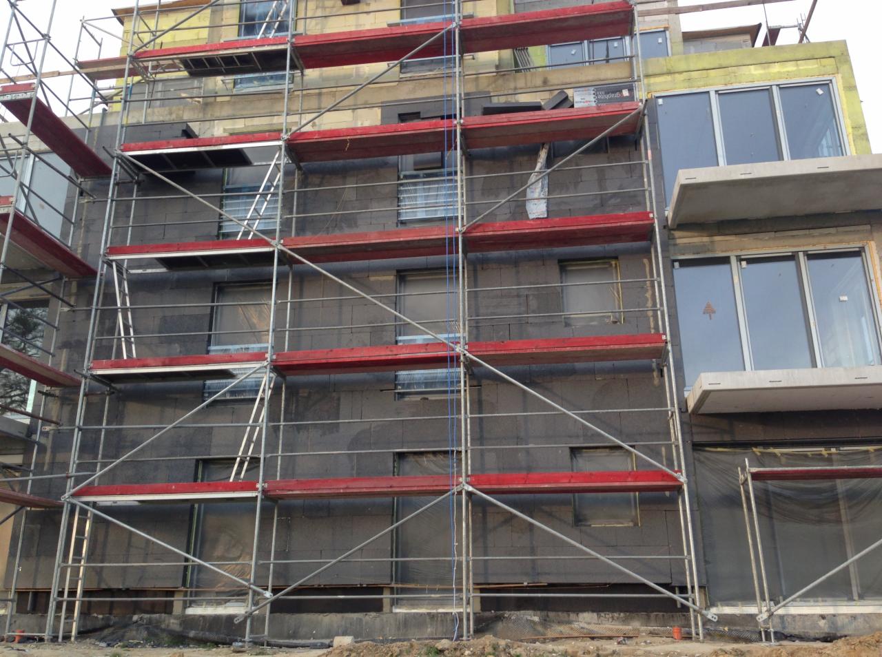 Ocieplanie elewacji budynku wielorodzinnego z użyciem styropianu grafitowego, widoczne rusztowania z czerwonymi poprzeczkami.