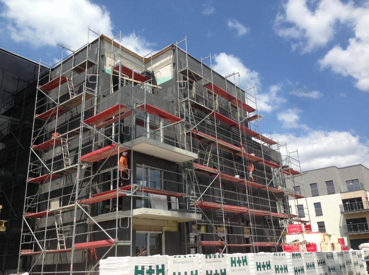 Wielopiętrowy budynek w trakcie ocieplania, pokryty rusztowaniami, z widocznymi pracownikami w pomarańczowych kamizelkach oraz paletami z bloczkami H+H na pierwszym planie.