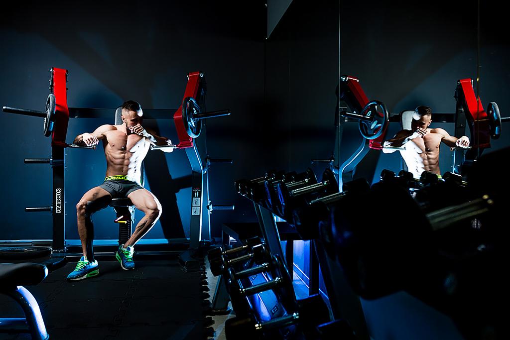 Mężczyzna bez koszulki siedzi na maszynie do ćwiczeń w siłowni, z widocznymi ciężarami i odbiciem w lustrze, oświetlony niebieskim światłem.