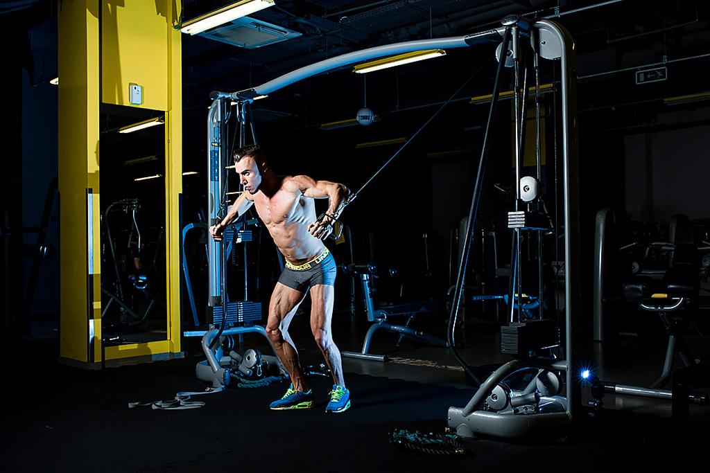 Mężczyzna bez koszulki ćwiczy na maszynie z obciążeniem w siłowni, widoczne mięśnie i napięcie, niebieskie buty sportowe.