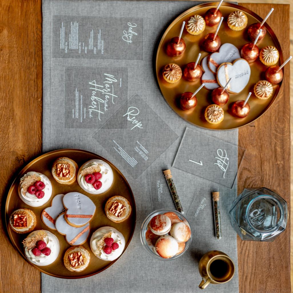 Dekoracje stołu weselnego z ciastkami, lizakami, makaronikami, herbatą w probówkach i przezroczystymi kartami z zaproszeniami i numerami stolików, ułożone na szarym bieżniku na drewnianym stole...