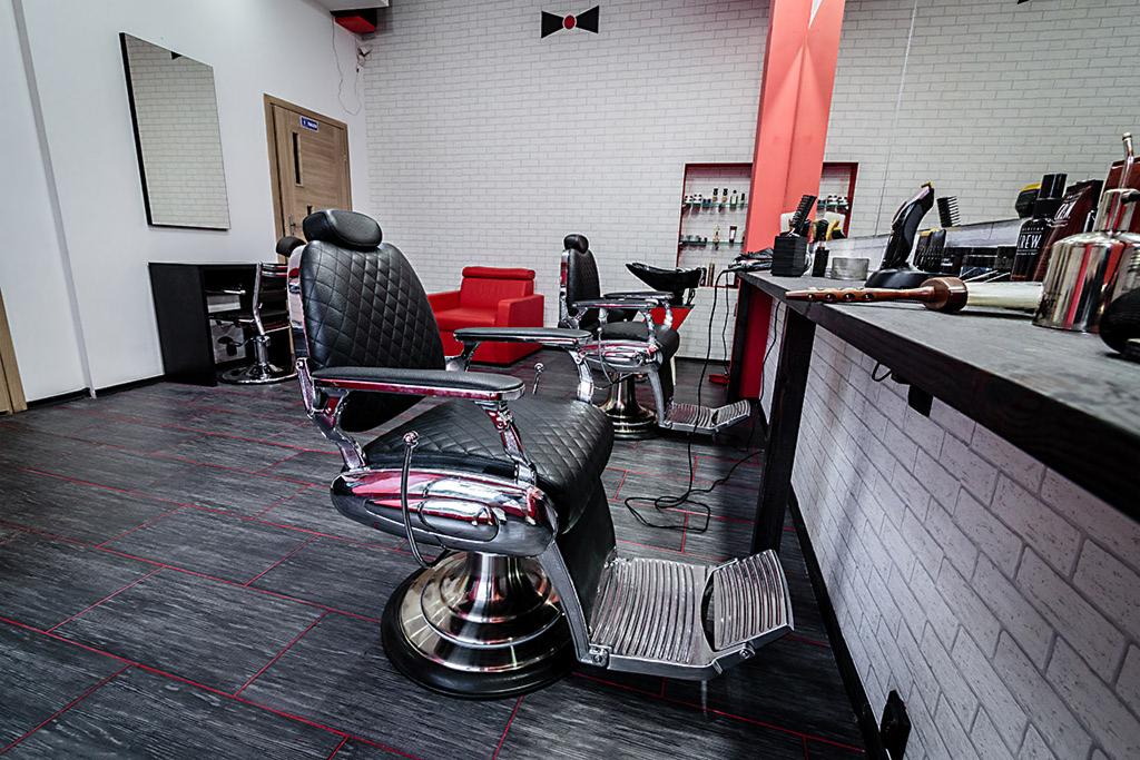 Wnętrze salonu barberskiego z dwoma fotelami fryzjerskimi z czarnej skóry i chromowanymi elementami, biała ściana z cegieł i czarną muchą jako dekoracją, ciemna podłoga z czerwonymi liniami.