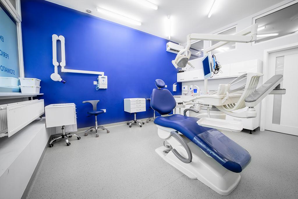 Nowoczesny gabinet dentystyczny z niebieską ścianą, białym sprzętem medycznym i fotelem dentystycznym z niebieskim obiciem.