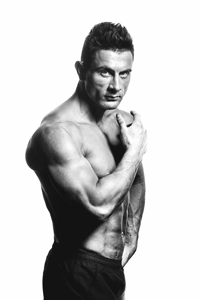 Czarno-białe, portretowe ujęcie muskularnego mężczyzny w sportowych spodenkach, pozującego z ręką na ramieniu, wyraźne kontrasty podkreślają definicję mięśni.