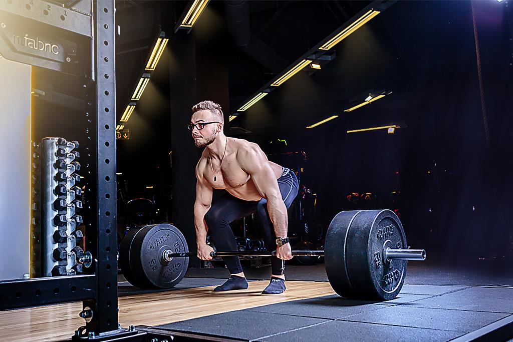 Mężczyzna w okularach, z odsłoniętą klatką piersiową, wykonuje martwy ciąg w siłowni, widoczne obciążenia i stojak na hantle.