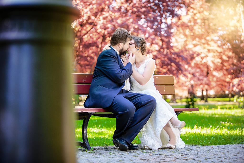 Nowożeńcy w objęciach siedzą na ławce w parku, panna młoda w koronkowej sukni i beżowych szpilkach, pan młody w granatowym garniturze, drzewa z różowymi kwiatami w tle.