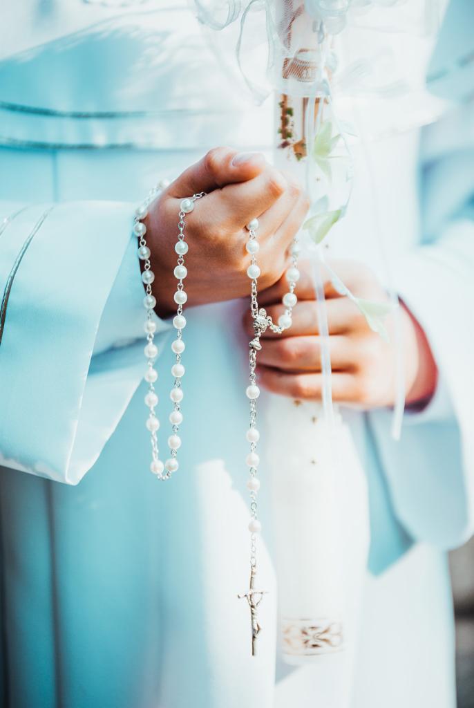 Dziecko w białej albie trzyma w dłoniach różaniec z pereł oraz świecę komunijną ozdobioną białą wstążką.