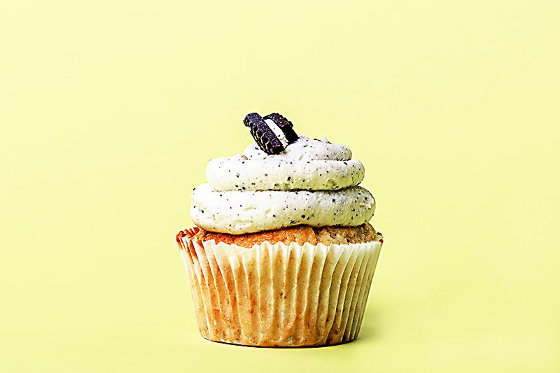 Babeczka waniliowa z kremem Oreo na żółtym tle, widoczne drobne czarne kropki w kremie i połówka ciastka na wierzchu.
