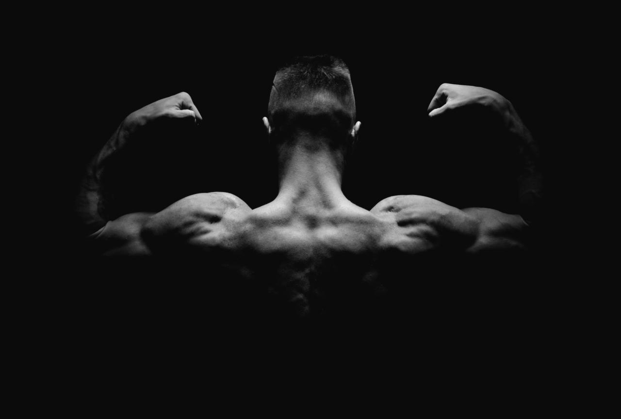 Czarno-białe zdjęcie od tyłu mężczyzny z napiętymi mięśniami ramion i pleców, oświetlonego z góry, na ciemnym tle.
