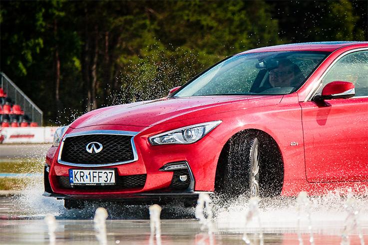 Czerwony samochód Infiniti z krakowską rejestracją KR 1FF37 dynamicznie przejeżdża przez wodny tor przeszkód, rozpryskując wodę, z widocznym kierowcą i trybunami w tle.