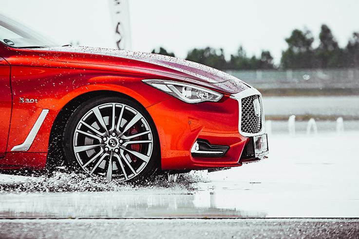 Czerwony samochód Infiniti Q60 z aluminiowymi felgami, dynamicznie przecinający kałużę, z widocznymi rozpryskami wody na karoserii i oponach.