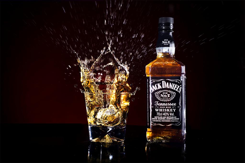 Butelka Jack Daniel's obok szklanki z lodem, z której dynamicznie pryska whiskey, na czarnym, błyszczącym blacie i ciemnym tle.