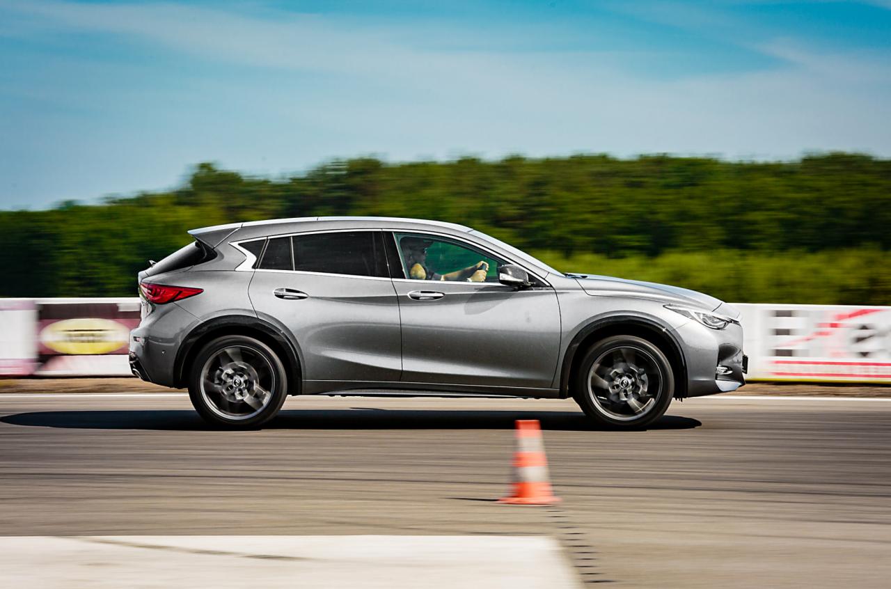 Srebrny crossover Infiniti QX30 w ruchu na torze wyścigowym, z pomarańczowym pachołkiem i zielonymi drzewami w tle.