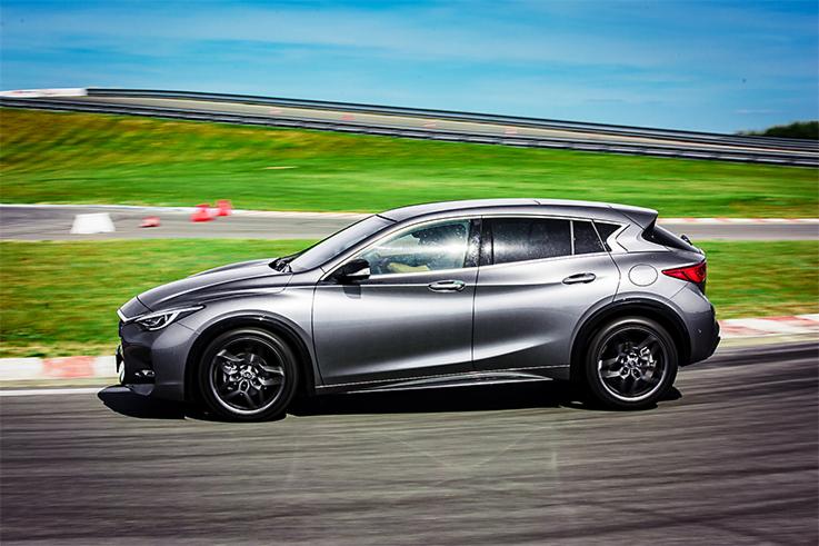 Szary crossover Infiniti QX30 na torze wyścigowym z zielonym wzgórzem i niebieskim niebem w tle, dynamiczne ujęcie w ruchu.