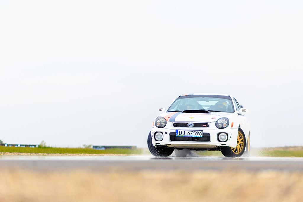 Biały samochód rajdowy Subaru Impreza z numerem startowym DJ 67596 na mokrej nawierzchni toru, widok z przodu, złote felgi, lekki deszcz.