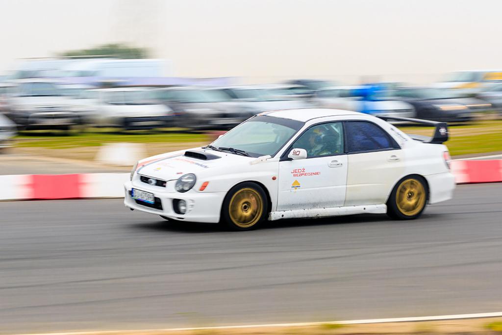 Biały samochód sportowy z czarnym spoilerem i złotymi felgami, pędzący po torze wyścigowym z rozmytym tłem innych pojazdów.