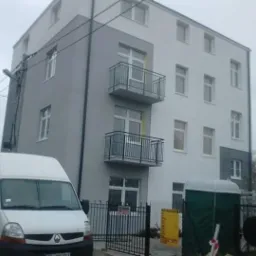 Odnowiony budynek wielorodzinny z balkonami, pomalowany na szaro i biało, z zaparkowanym białym vanem i przenośną toaletą na pierwszym planie, widok z niskiej perspektywy w pochmurny dzień.