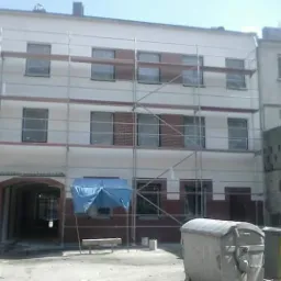 Budynek w trakcie renowacji elewacji z widocznymi rusztowaniami, częściowo otynkowaną fasadą i elementami ceglanymi, zabezpieczonym wejściem oraz kontenerami na gruz w tle.