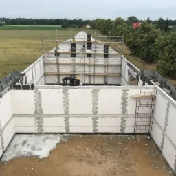 Wznoszenie ścian parteru domu z białych bloczków na fundamencie, widoczne rusztowania i częściowo zagospodarowany teren budowy z zewnątrz.