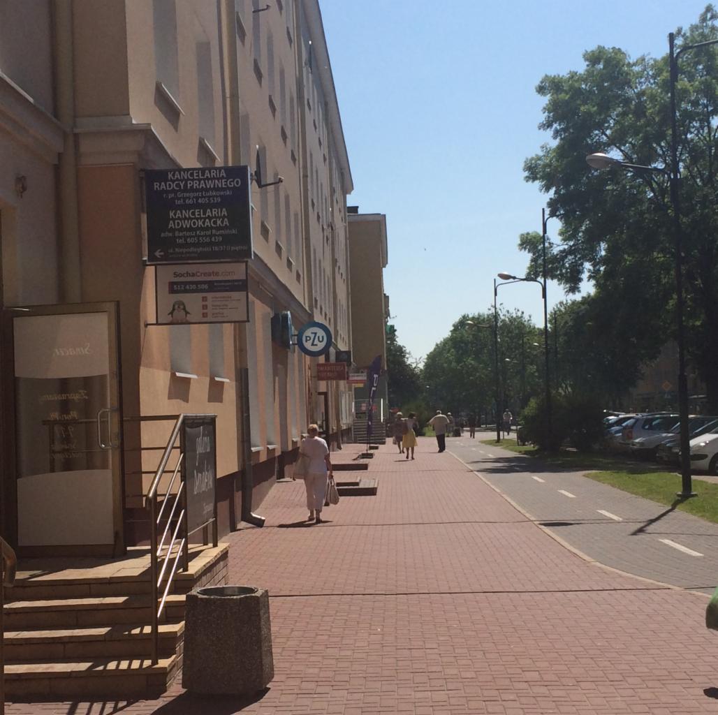 Budynek z szyldami Kancelarii Radcy Prawnego i Kancelarii Adwokackiej, widok z chodnika wyłożonego czerwoną kostką, w tle spacerujący ludzie i zaparkowane samochody.