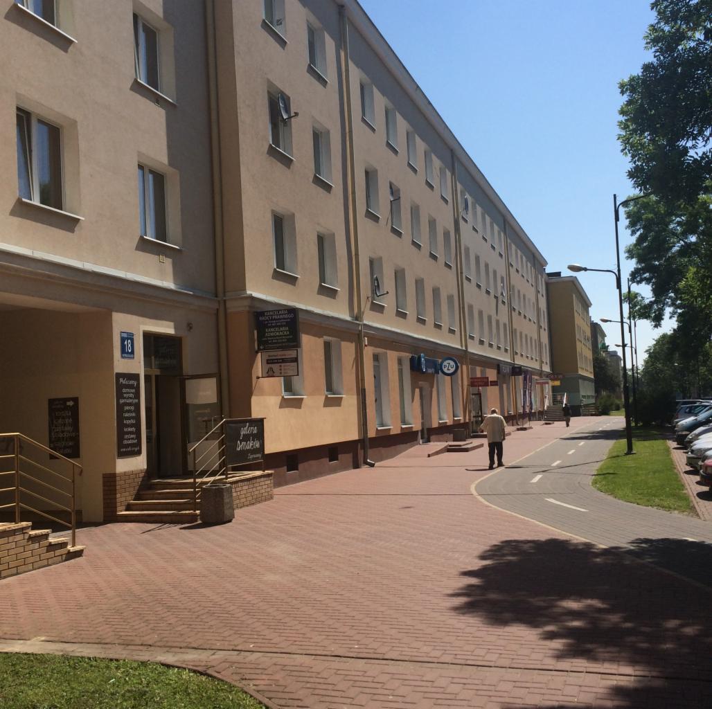 Fasada budynku z biurami, w tym Kancelaria Radcy Prawnego, z chodnikiem z czerwonej kostki i osobą idącą wzdłuż niego w słoneczny dzień.