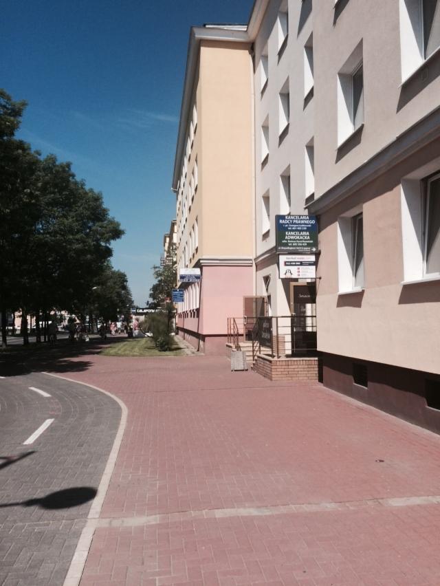 Budynek z szyldem informującym o Kancelarii Radcy Prawnego i Adwokata, widoczny z poziomu chodnika wyłożonego cegłą.