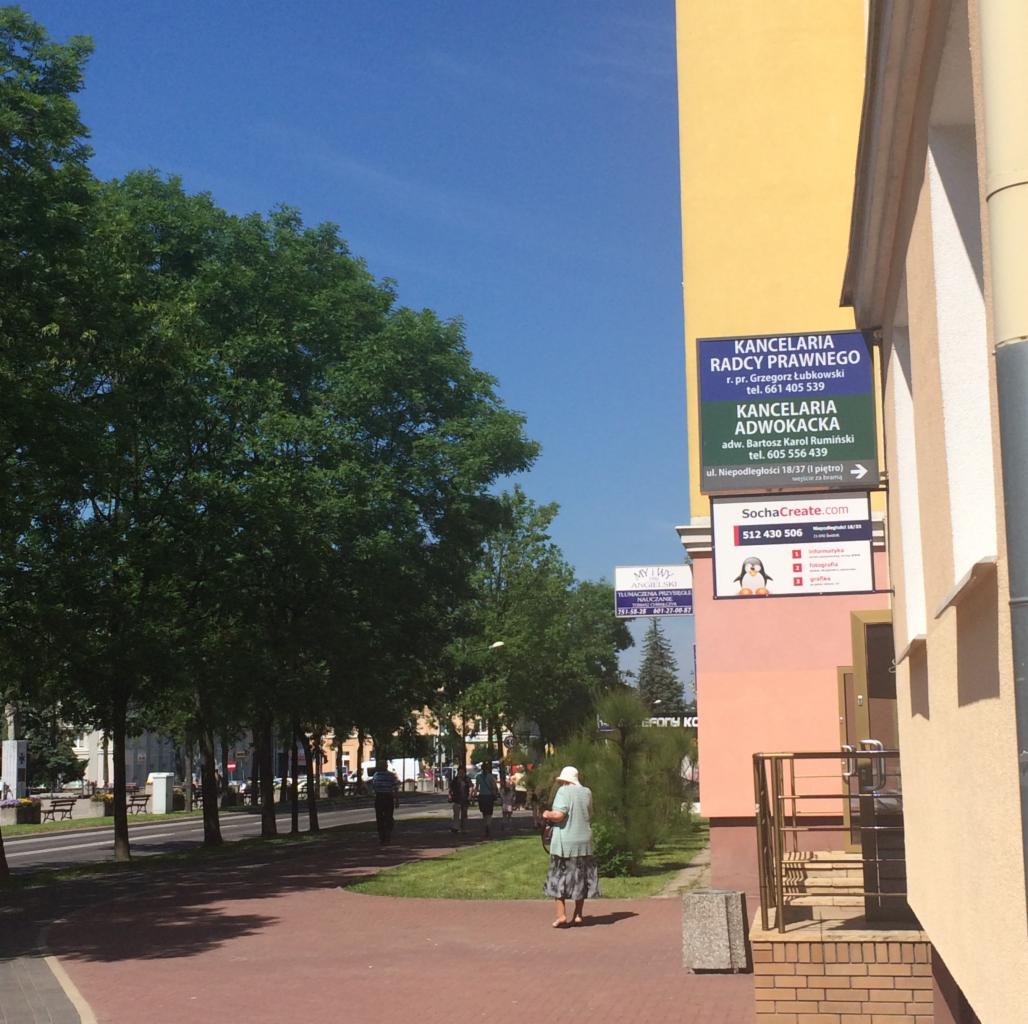 Tablica informacyjna z nazwą Kancelarii Radcy Prawnego i Kancelarii Adwokackiej na elewacji budynku, widok z chodnika z przechodzącą osobą.