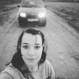 Czarno-białe selfie kobiety na polnej drodze z samochodem Fiat Cinquecento w tle, oświetlonymi reflektorami i widoczną tablicą rejestracyjną.