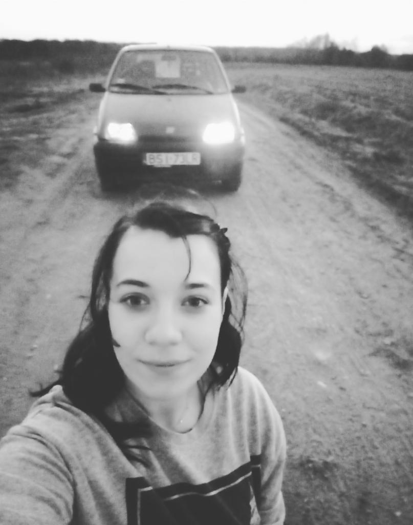 Czarno-białe selfie kobiety na polnej drodze z samochodem Fiat Cinquecento w tle, oświetlonymi reflektorami i widoczną tablicą rejestracyjną.