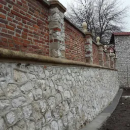 Fragment muru z czerwonej cegły i jasnego kamienia, zwieńczonego kamiennymi kolumnami z kulami, widok z dołu.