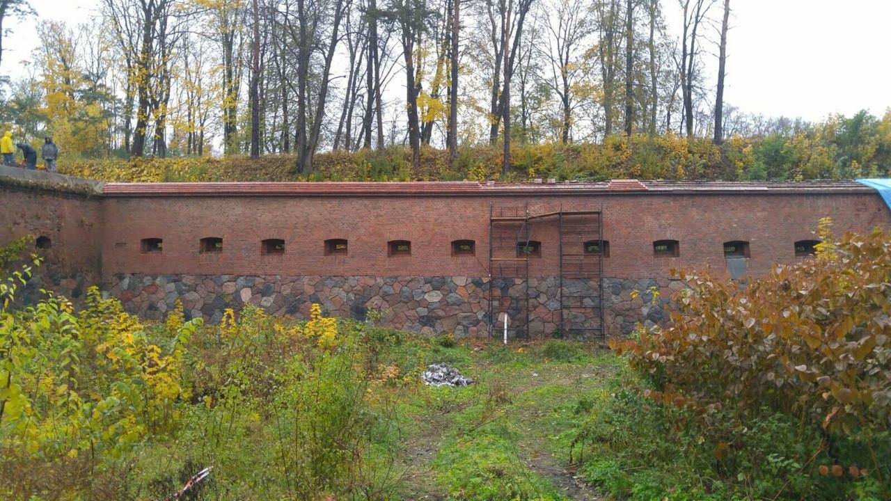 Fragment muru obronnego z cegły i kamienia z małymi oknami strzelniczymi, zarośnięty roślinnością, z rusztowaniem po prawej stronie i osobami na szczycie muru w oddali.