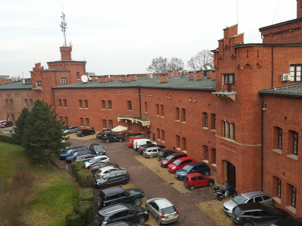 Widok z góry na duży, ceglany budynek z wieżą i anteną, z parkingiem pełnym samochodów na dziedzińcu. Budynek w stylu historycznym, prawdopodobnie odrestaurowany.