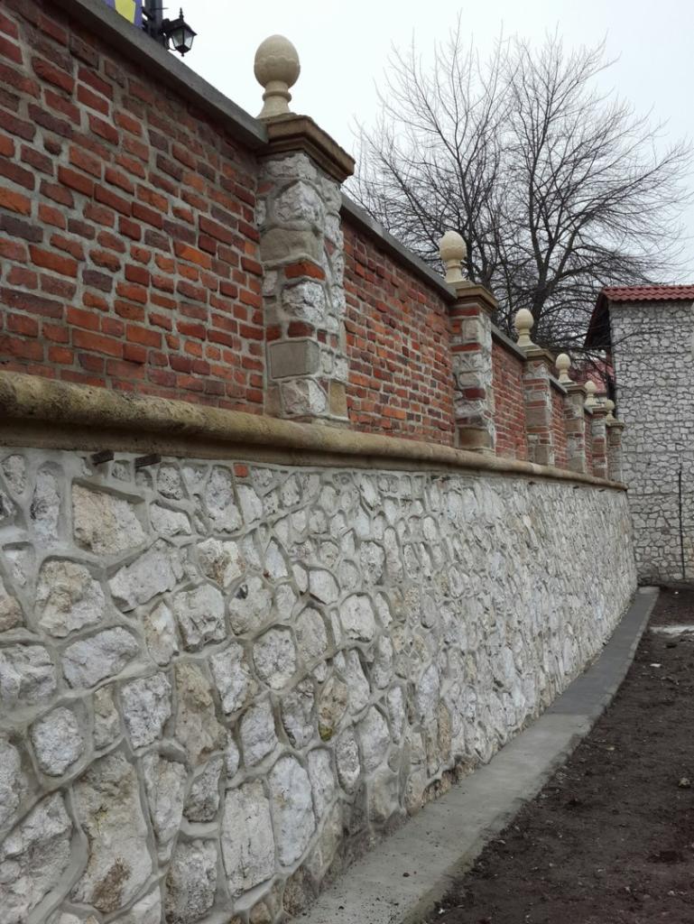 Fragment muru z czerwonej cegły i jasnego kamienia, zwieńczonego kamiennymi kolumnami z kulami, widok z dołu.