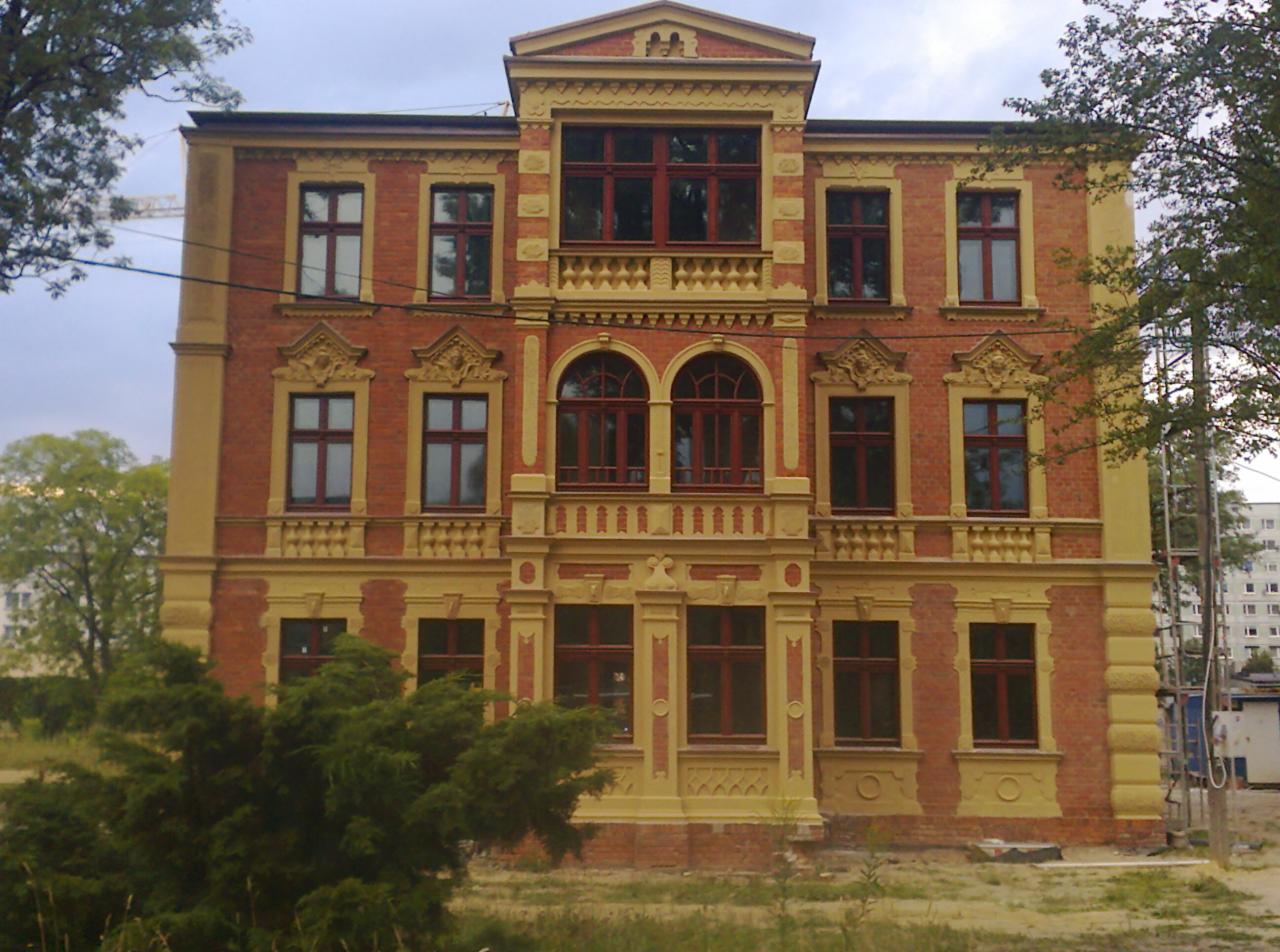 Odrestaurowana, trzypiętrowa kamienica z czerwonej cegły z żółtymi zdobieniami, widoczne elementy architektoniczne i dekoracyjne, w tle drzewa i budynek mieszkalny.