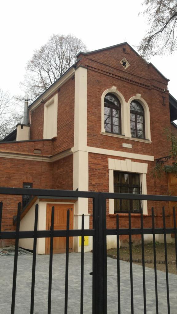 Odrestaurowana, dwupiętrowa kamienica z czerwonej cegły, z charakterystycznymi łukowymi oknami i jasnymi, kremowymi elementami dekoracyjnymi, widziana zza czarnego, metalowego ogrodzenia.