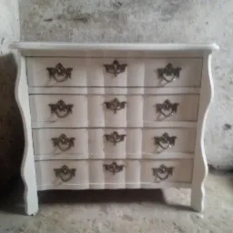 Biała komoda z trzema szufladami i ozdobnymi, metalowymi uchwytami na tle surowej ściany.