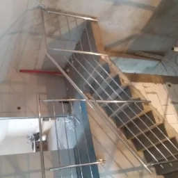 Montaż metalowej balustrady na betonowych schodach w surowym wnętrzu, widoczne przewody i niedokończone wykończenie.