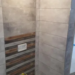 Ściana łazienki wyłożona szarymi płytkami imitującymi beton, z wstawką dekoracyjną z płytek drewnopodobnych, widoczne żółte zaślepki na rurach i częściowo zamontowany przycisk spłuczki.