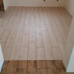 Położona jasna podłoga drewnopodobna w pokoju, kontrastująca z ciemniejszym fragmentem podłogi przy wejściu, widoczne puszki elektryczne na ścianie.