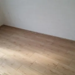 Ukończona podłoga z płytek imitujących drewno w rogu pomieszczenia, widoczna puszka elektryczna na ścianie.