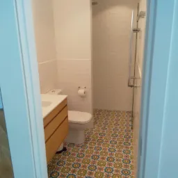 Wnętrze łazienki po remoncie: wzorzysta mozaika na podłodze, białe płytki na ścianach, szafka pod umywalką z jasnego drewna i kabina prysznicowa ze szklanymi drzwiami.