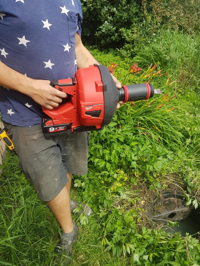 Mężczyzna trzyma w rękach elektryczną przepychaczkę do rur Milwaukee 9.0 Ah, przygotowując się do pracy przy studzience kanalizacyjnej na zewnątrz, w otoczeniu zieleni.