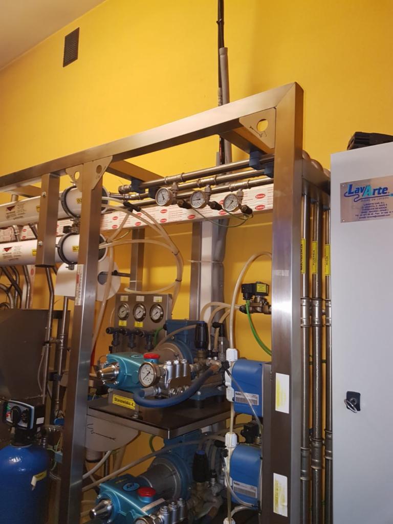 Kompleksowa instalacja przemysłowa z licznymi rurami, manometrami i pompami na tle żółtej ściany, widoczny fragment panelu sterującego.