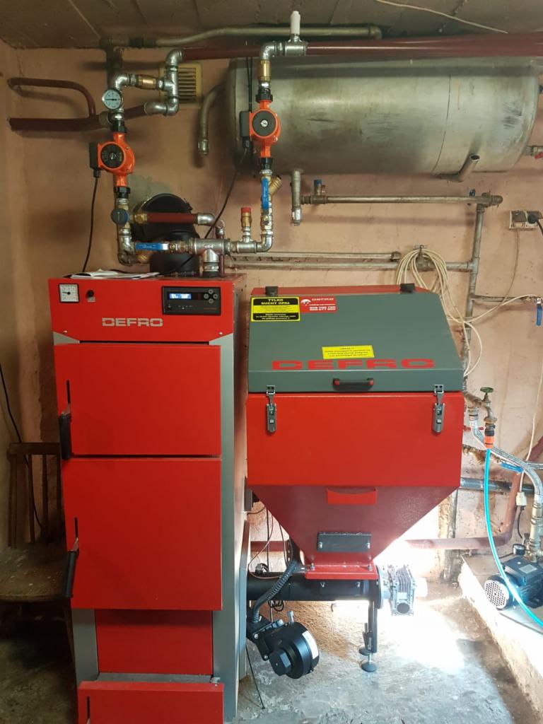 Czerwony piec DEFRO z podajnikiem, zbiornik na wodę i instalacja grzewcza z pompami w kotłowni o nierównych ścianach.