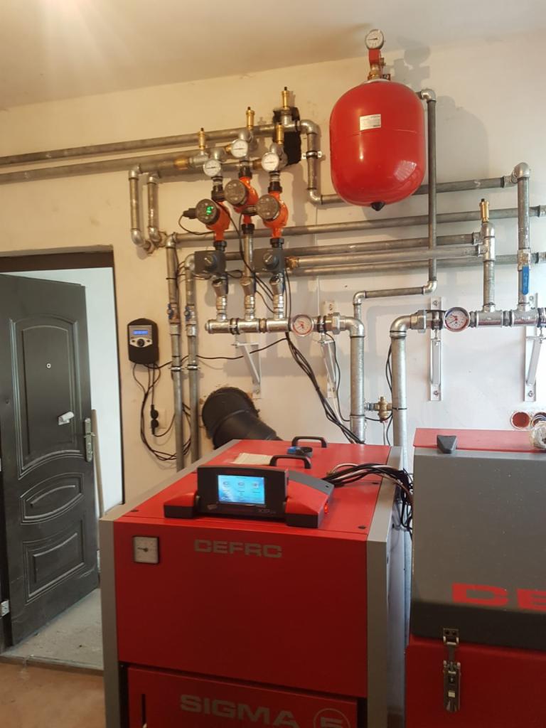 Kotłownia z kotłem na paliwo stałe Defro Sigma, zbiornikiem wyrównawczym i rozbudowaną instalacją hydrauliczną z pompami obiegowymi i manometrami.
