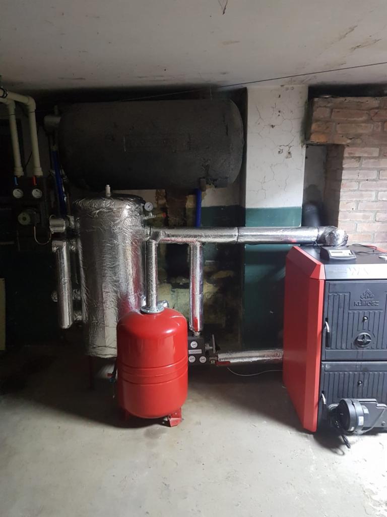 Kotłownia z kotłem na paliwo stałe, zbiornikiem akumulacyjnym owiniętym folią aluminiową, czerwonym naczyniem wzbiorczym i instalacją rurową w piwnicy z surowymi ścianami.