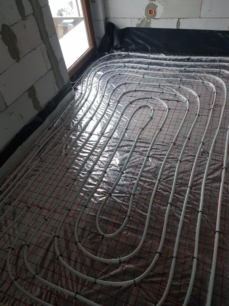 Równomiernie ułożone rury systemu ogrzewania podłogowego na folii z siatką, widoczne elementy mocujące, ściany z pustaków i okno w surowym stanie.