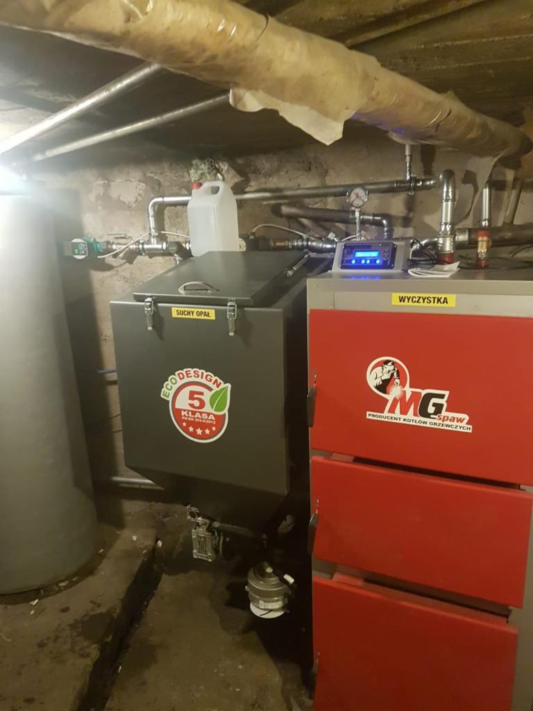 Widok kotłowni z dwoma kotłami: szarym z napisem 'Suchy Opał' i czerwonym z logo producenta 'MG Spaw'. Widoczne rury instalacji grzewczej i zbiornik buforowy. Na kotłach widoczne panele sterowania.