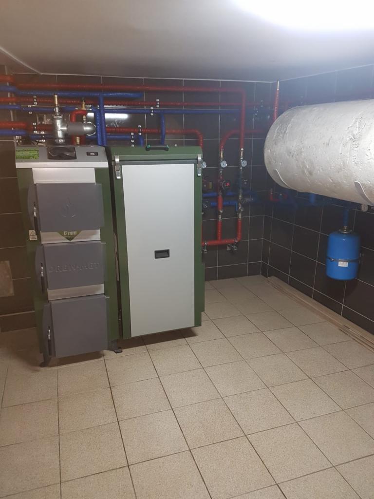 Kotłownia z kotłem na paliwo stałe marki DREWMET, zasobnikiem ciepłej wody i instalacją grzewczą z rur miedzianych i plastikowych w piwnicy wyłożonej płytkami ceramicznymi.