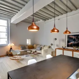 Jasne wnętrze z drewnianym sufitem, trzema miedzianymi lampami wiszącymi nad stołem, oraz narożną sofą w tle, oświetlone naturalnym światłem wpadającym przez duże okno.